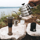 Person pouring coffee from a metal kettle into an AeroPress on a rock with a scenic background.